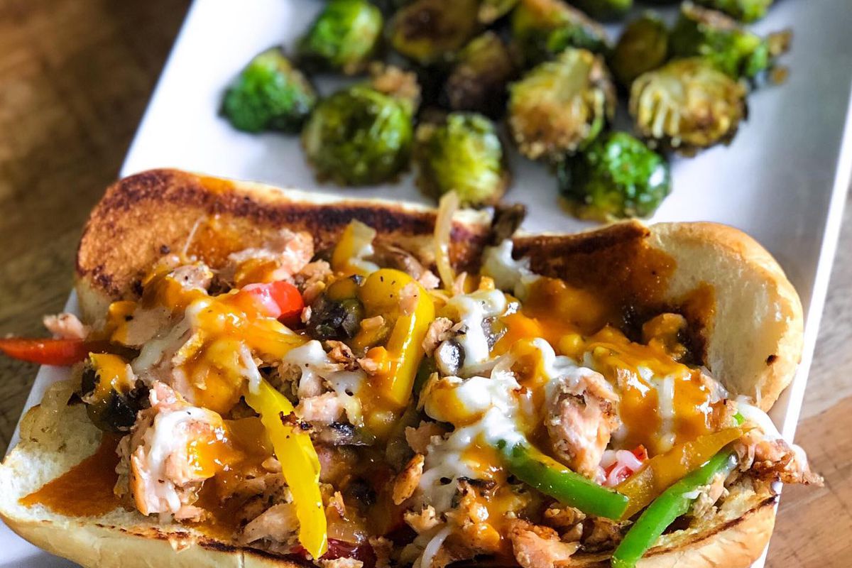 A white rectangular plate with a sandwich stuffed with grilled salmon, grilled trinity pepper mix, mushrooms, onions, and melted vegan mozzarella on brioche bread next to a side of roasted Brussels sprouts