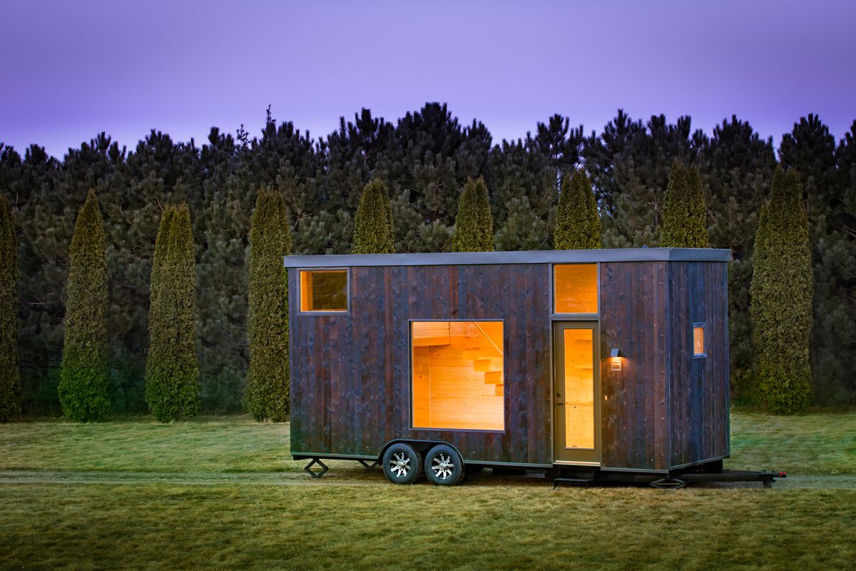 A rectilinear mobile unit clad in charred wood siding and punctuated with a large square window, glazed entryway and transom window, and other small windows sits in a field in front of a forest of trees. 