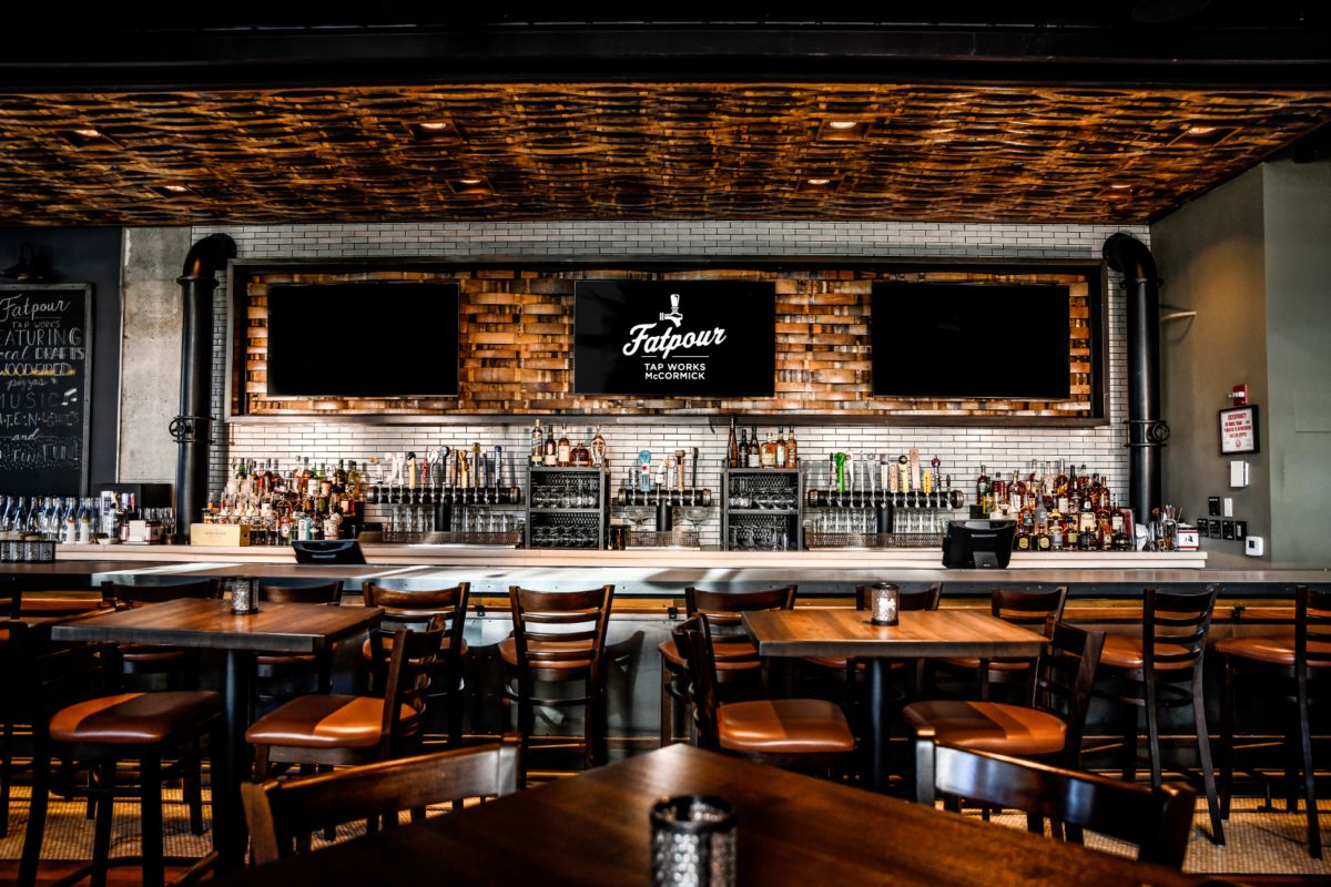 A large bar space is filled with dark wood tables and chairs. A tiled wall behind the bar offsets bottles, taps, and coolers. The brown ceiling appears to be wood woven into a basket-weave texture. Three TVs hang over the bar. 