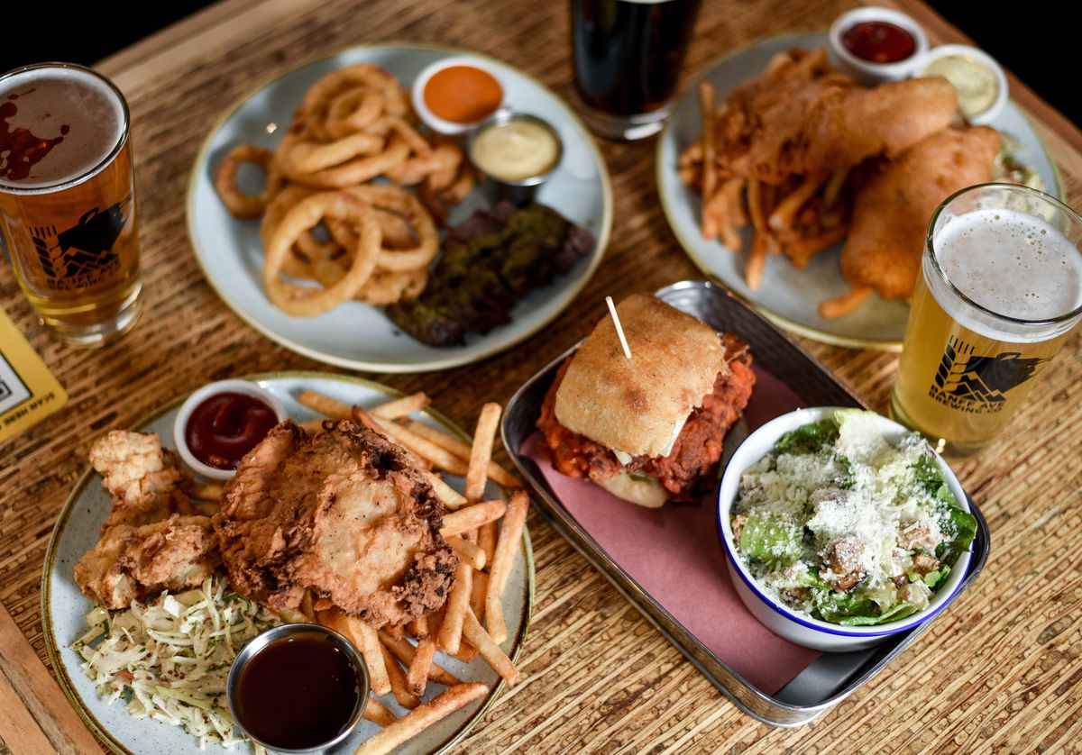 Plates of fried chicken, fried fish, onion rings, and fried chicken sandwich, with sides like slaw, chopped salad, and sauces, as well as a few pints of beer