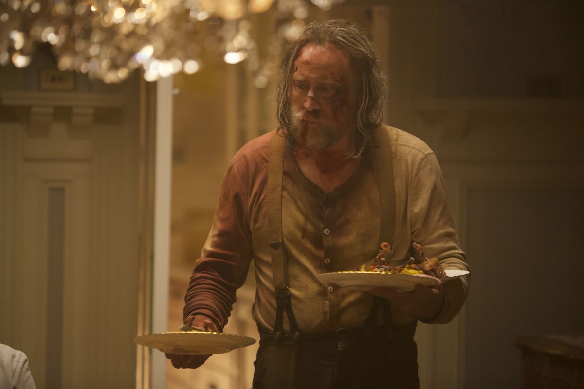 A still from the movie ‘Pig,’ featuring Nicolas Cage looking bruised and bloody, carring three plates of food in his arms.