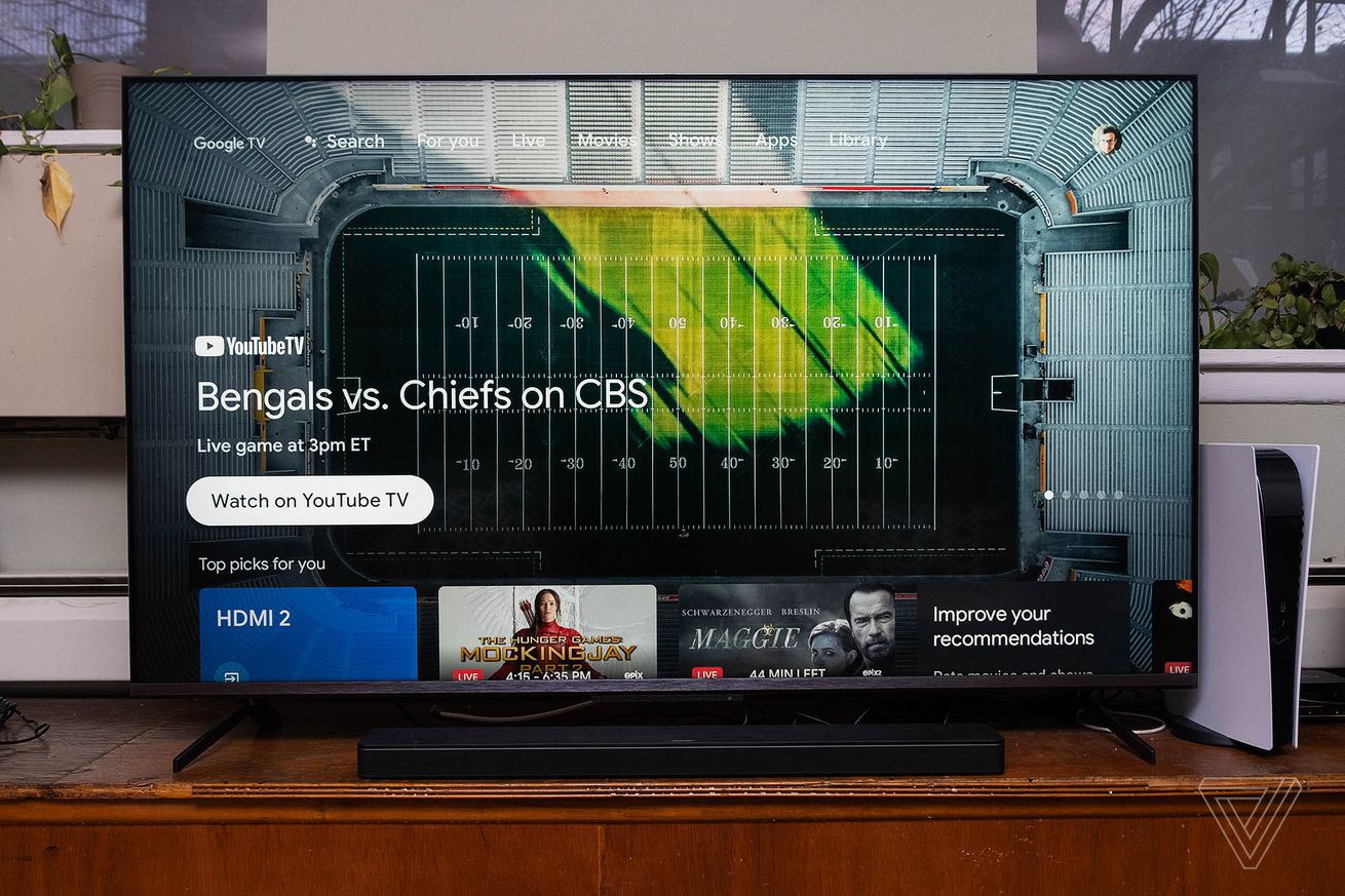 The TCL 6-Series TV standing on a piece of furniture beside a PlayStation 5, displaying the Google TV home screen.