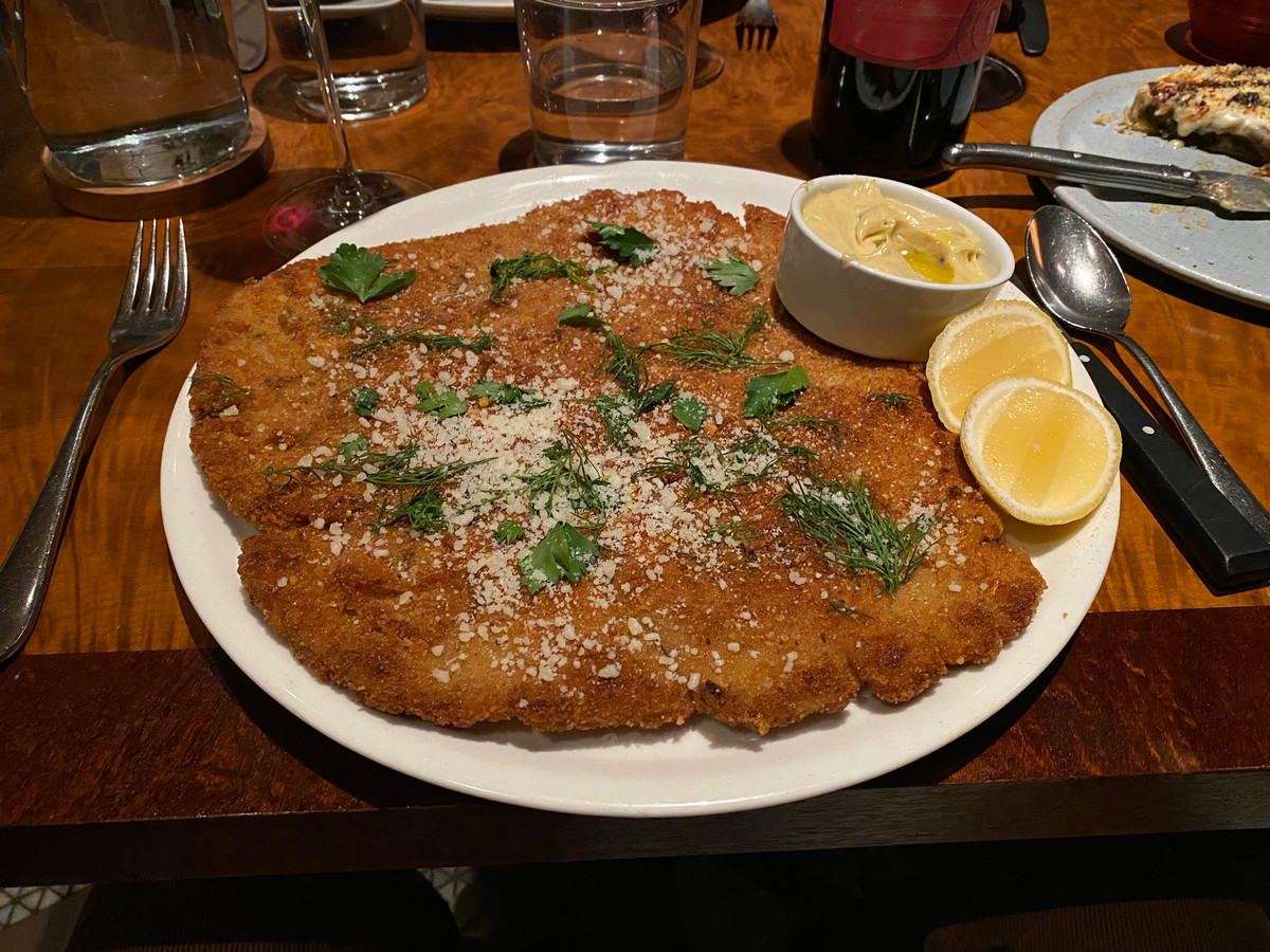 An oversized slab of pork Milanese on a plate is adorned with green herbs and wedges of lemon.