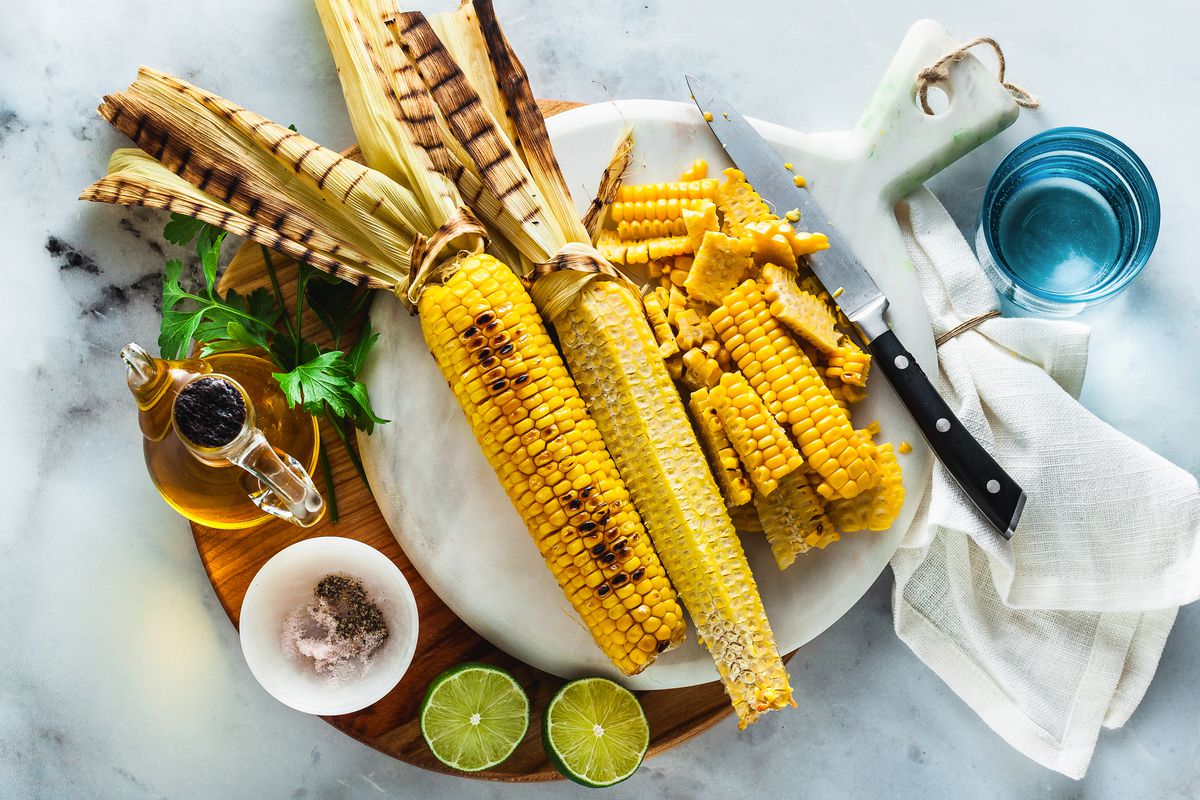 A plate of grilled corn on the cob surrounded by cut off wedges of kernels, limes, oil, and a blue glass on a white marble table top.