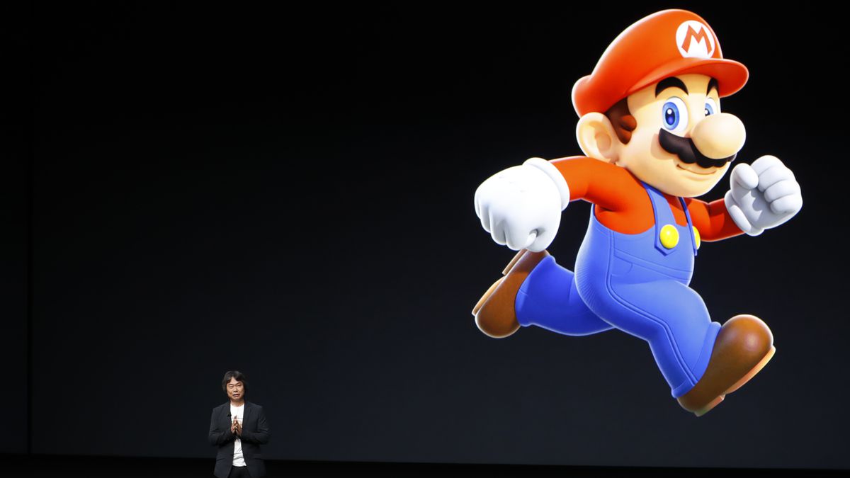 Photo of Shigeru Miyamoto speaking on a stage in front a large projection of Super Mario in the top right corner of the backdrop.