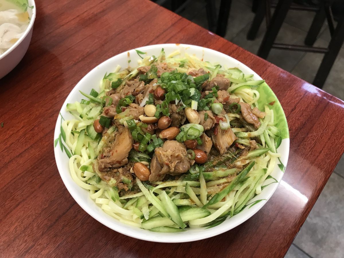 A bowl with cold noodles, spicy chicken, and cucumbers topped with scallions and peanuts sits on a wooden table.