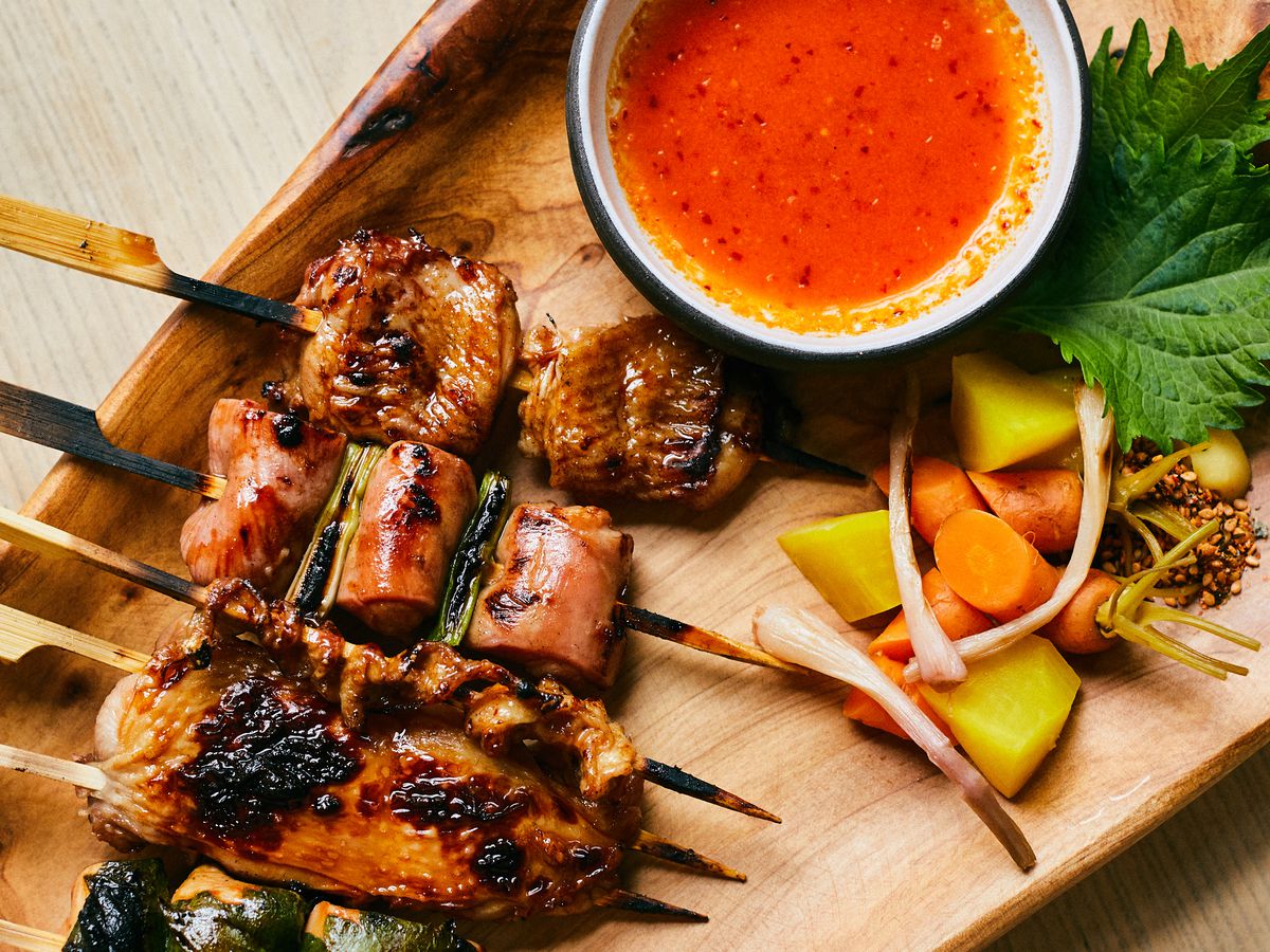 Skewers of meat next to pickled vegetables and red sauce on a wooden plate.