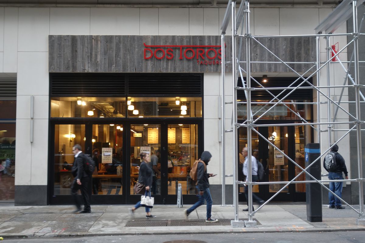 The gray exterior of a restaurant with scaffolding partially covering the building and people walking by outside.