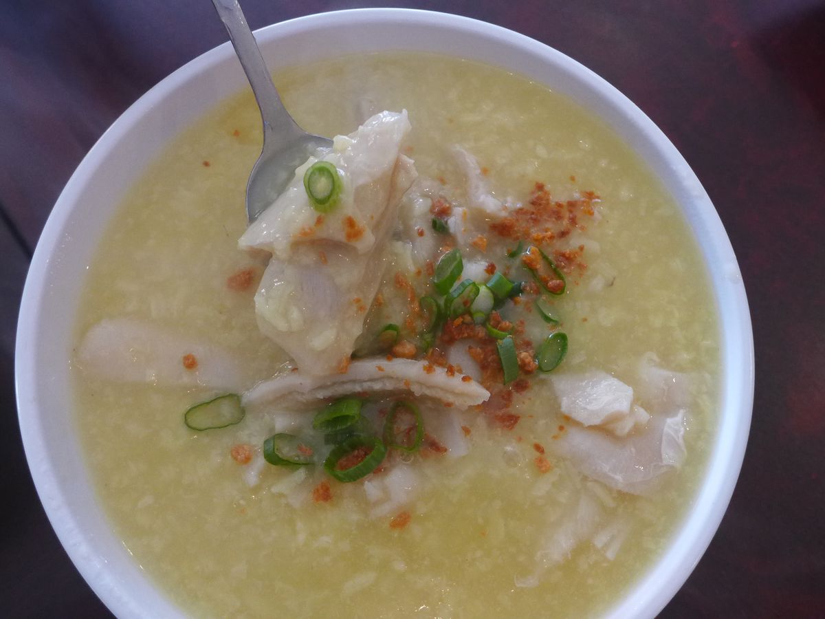 A yellowish rice soup with chunks of fish visible, one suspended on a spoon over the soup.