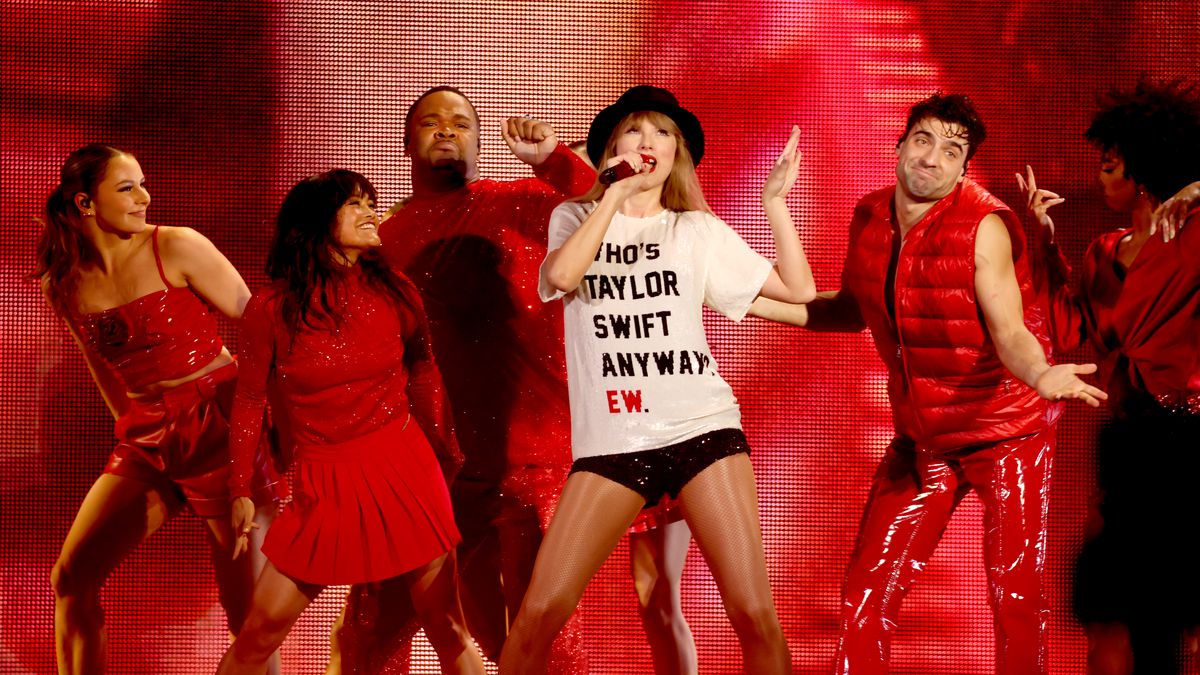 A singer in a white shirt that reads Who Is Taylor Swift Anyway? Ew. With dancers in red behind her.