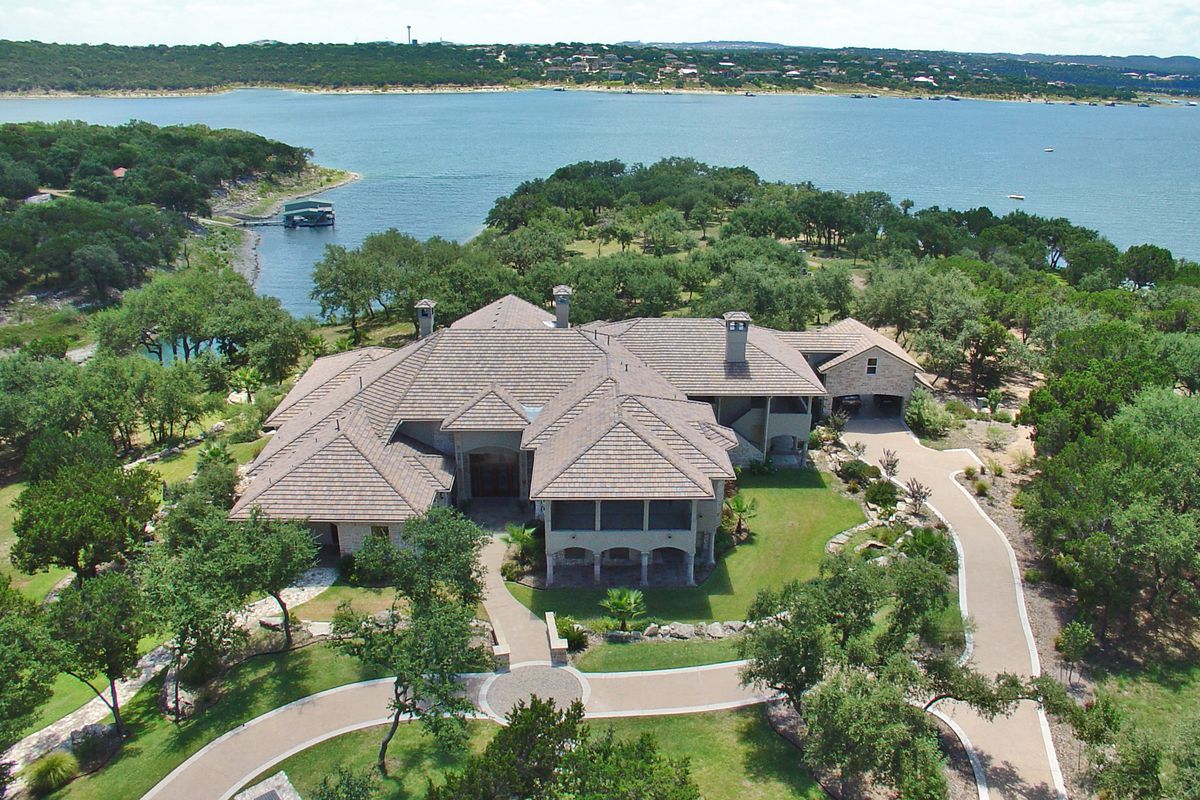 Aerial view of very large, two story mansion compound on green peninsula with lake in background