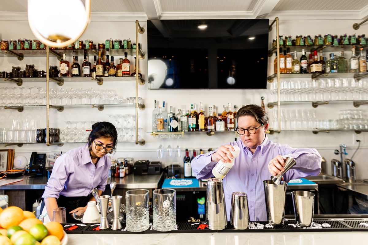 Two bartenders behind the bar at Restaurant Beatrice. One is making a cocktail in a shaker, the other is searching behind the bar.