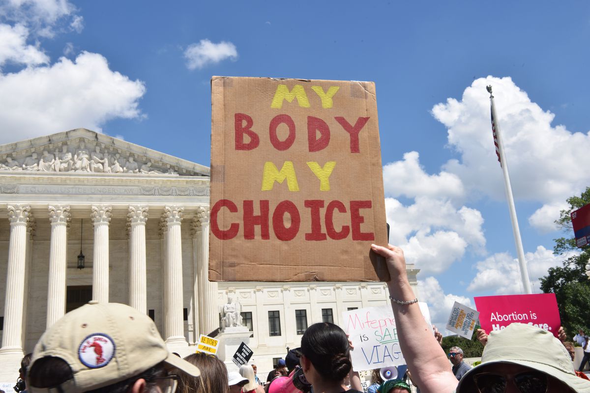 In front of the Supreme Court building’s white marble columns and steps, a crowd of people hold signs. The center of the photo shows a brown cardboard sign held above a marcher’s head that reads “My body my choice.”