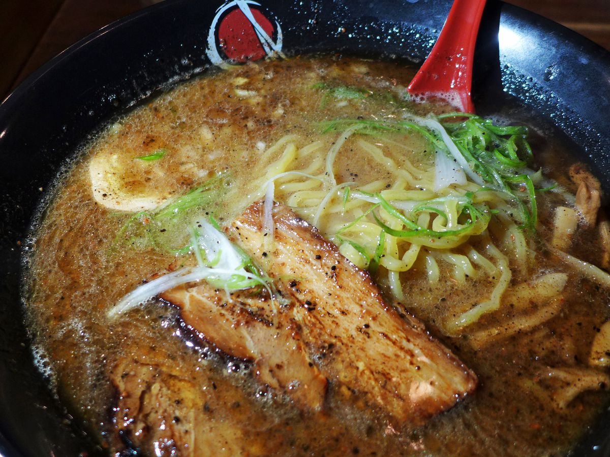 A tangle of ramen noodles with pork chashu in a black bowl