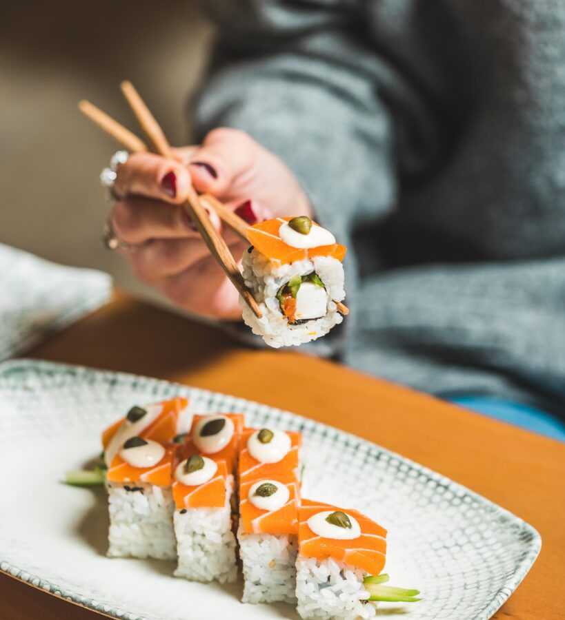 A diner holds a piece of plant sushi roll with chopsticks above a plate.