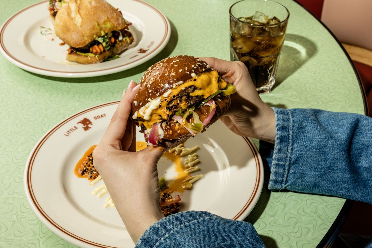 Two hands grabbing a burger off a plate on a table.