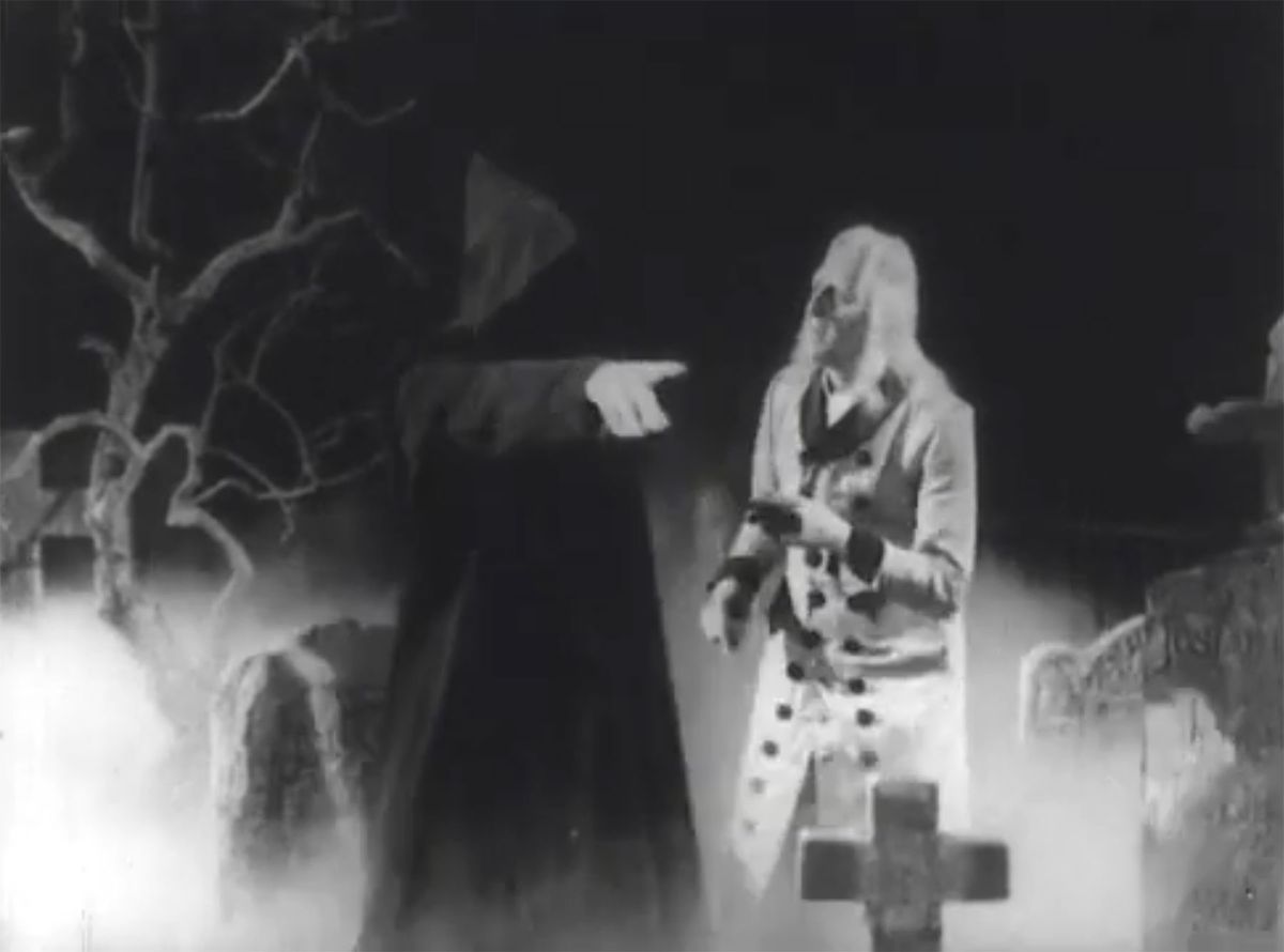 A robed ghost points offscreen in a cemetery to direct a scrooge with long white hair