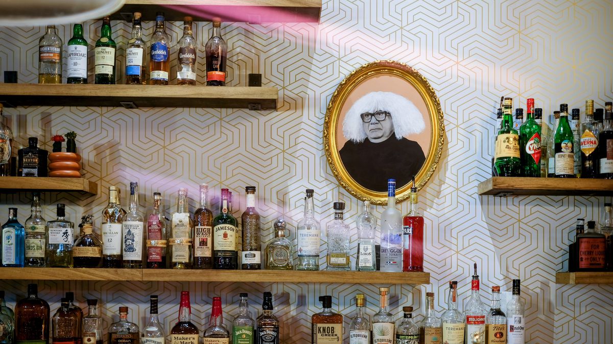 A gold-framed painting of Danny Devito wearing a white wig on a wall loaded up with shelves and liquor bottles.