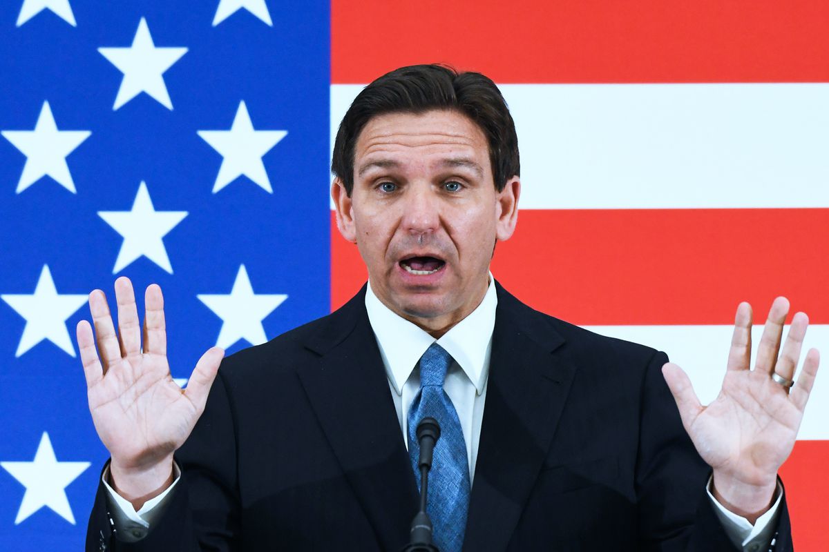 Ron DeSantis, wearing a blue suit, gestures with both hands open while speaking, in front of a large US flag.