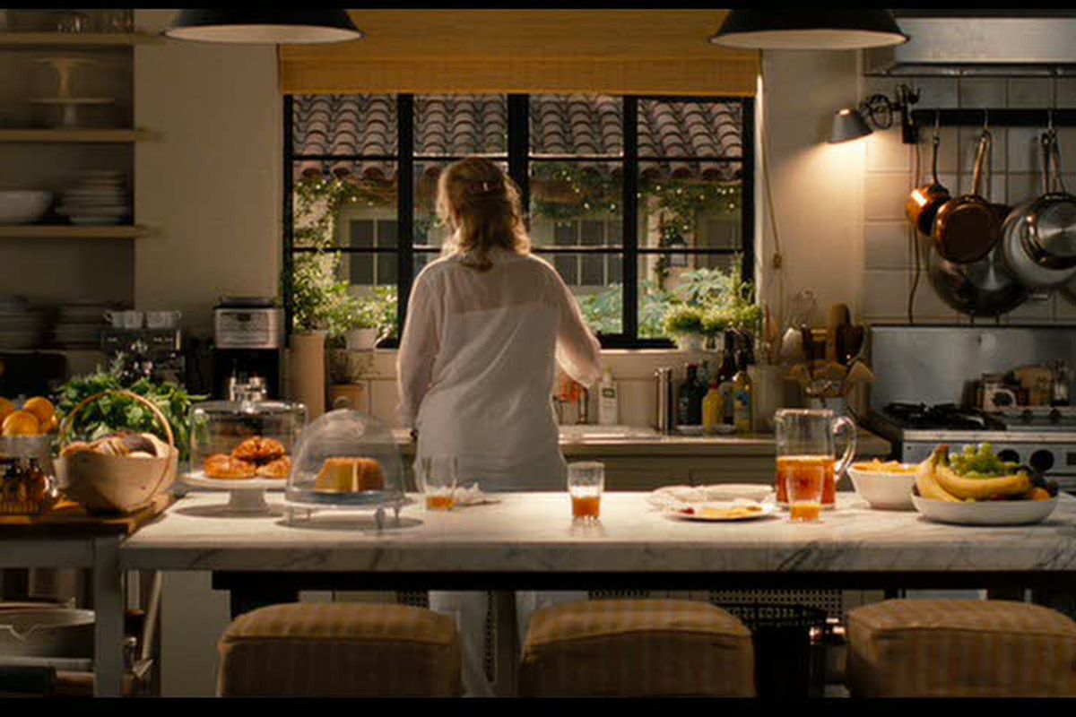 A woman with her back turned to the sink in front of a large window in a beautiful rustic kitchen. 