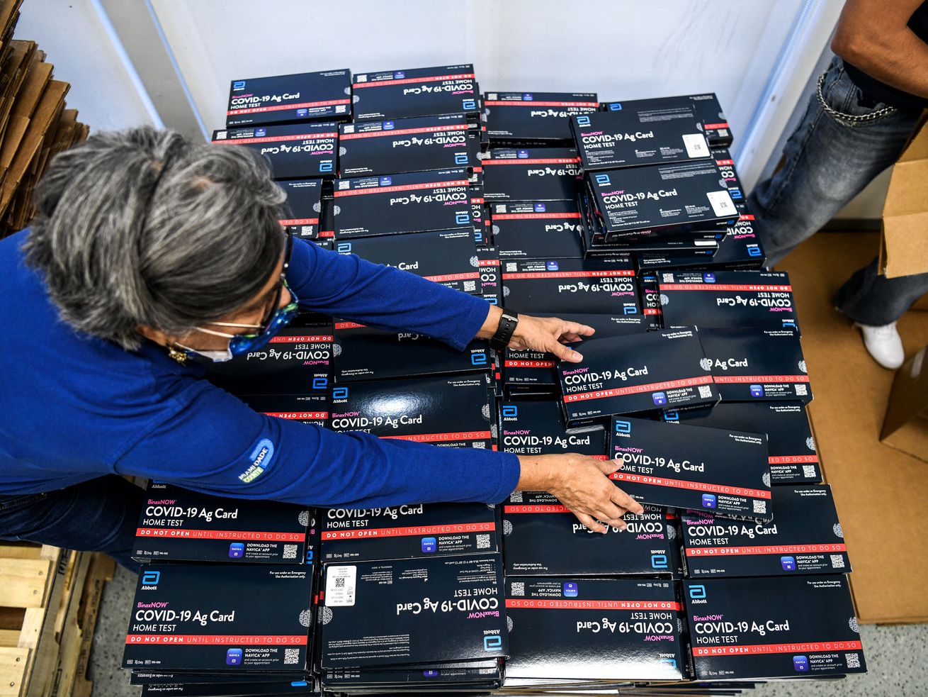 A person reaching across a large number of boxes of Covid-19 tests.