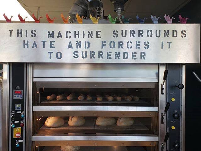 A large commercial oven filled with bread. On the top of the oven it says, “This machine surrounds hate and forces it to surrender.”