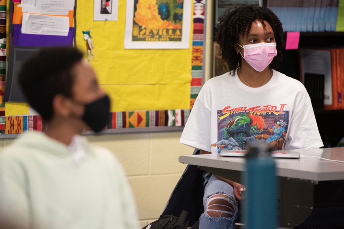 Students sit in a Chicago classroom, both with a mask on and a yellow bulletin board behind them. One student is in the foreground but blurry, the student in the background is sharper in the image. 