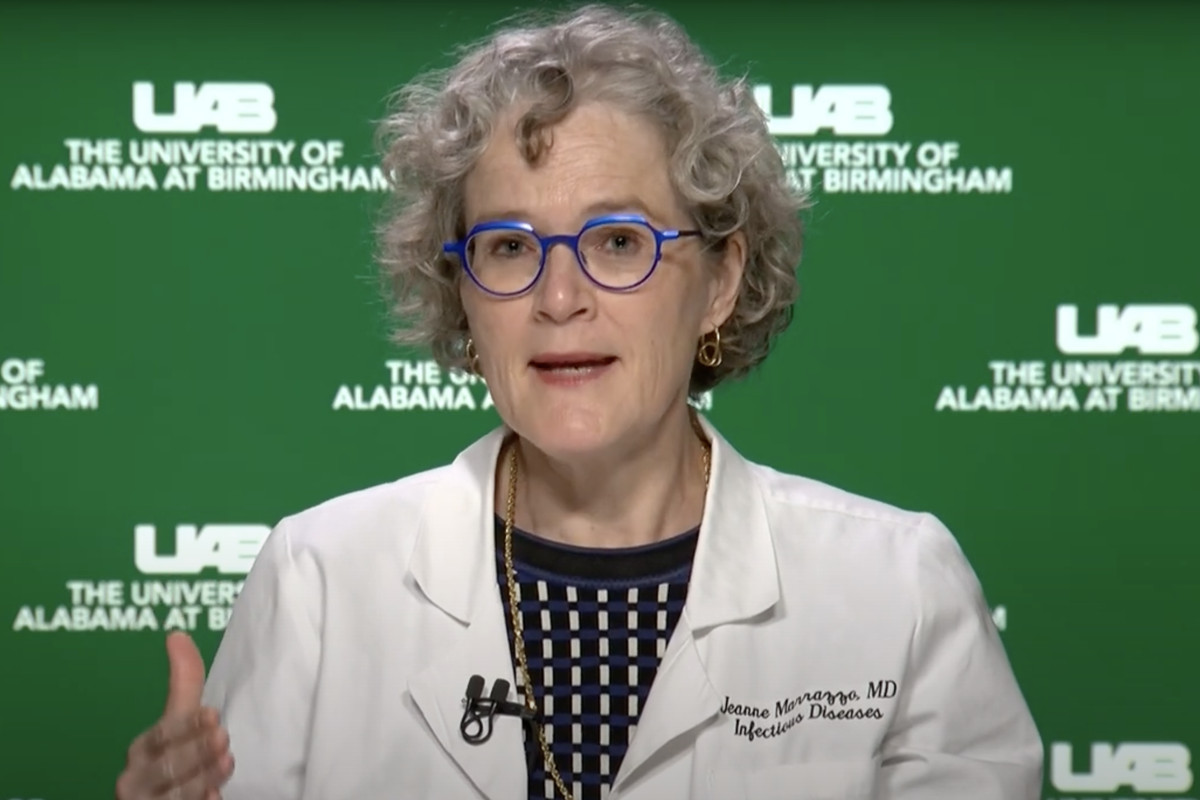 A white woman with glasses speaks against a green and white backdrop.