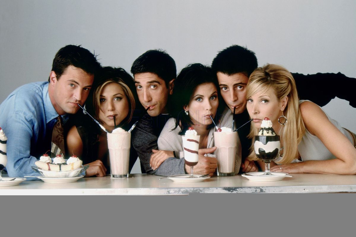 The cast of “Friends” leaning forward and sipping on milkshakes sitting on a table.