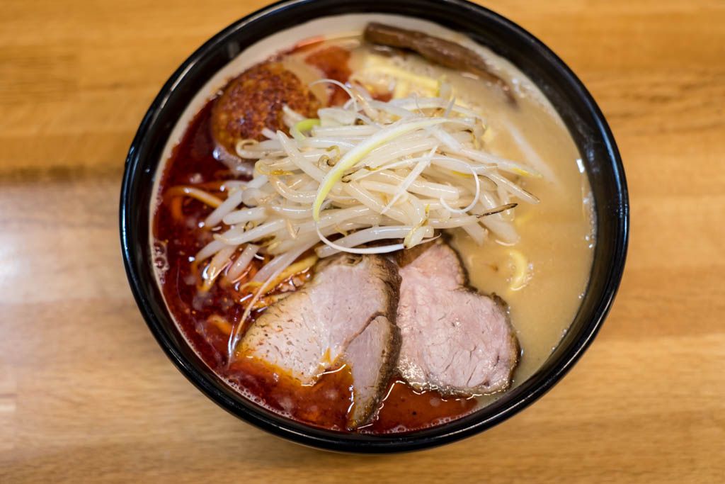 From above, a bowl of ramen with two large slices of pork, sprouts, deep red broth on one side and pale yellow on the other, on a wooden countertop.