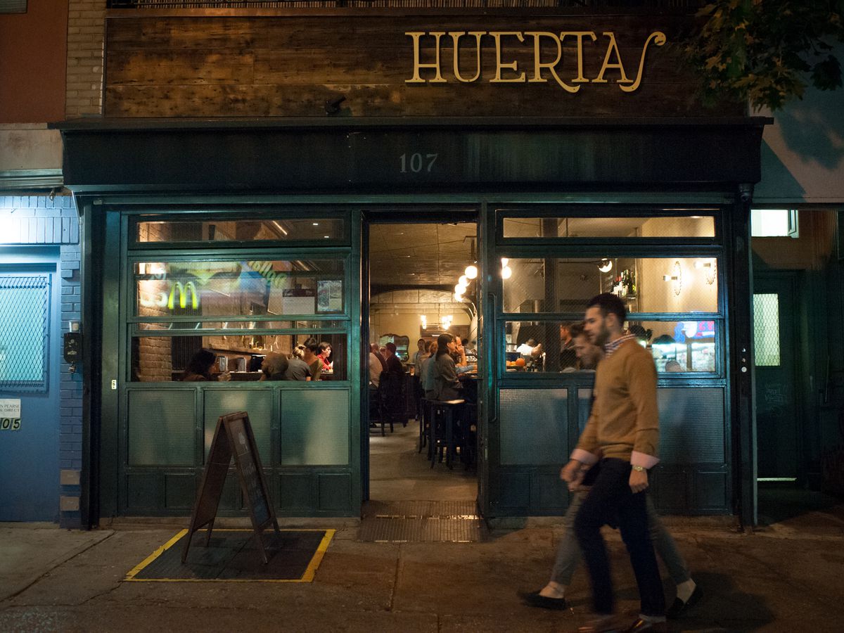 The Huertas restaurant storefront is filled with people, as two people walk by at night.