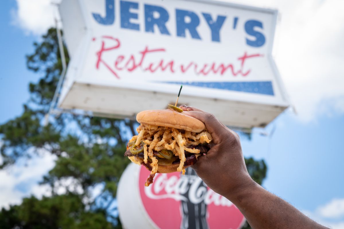 A man’s hand holds up a burger that is pinned together with a toothpick and over stuffed with fried onions. Behind it is a sign that reads, “Jerry’s Restaurant.”