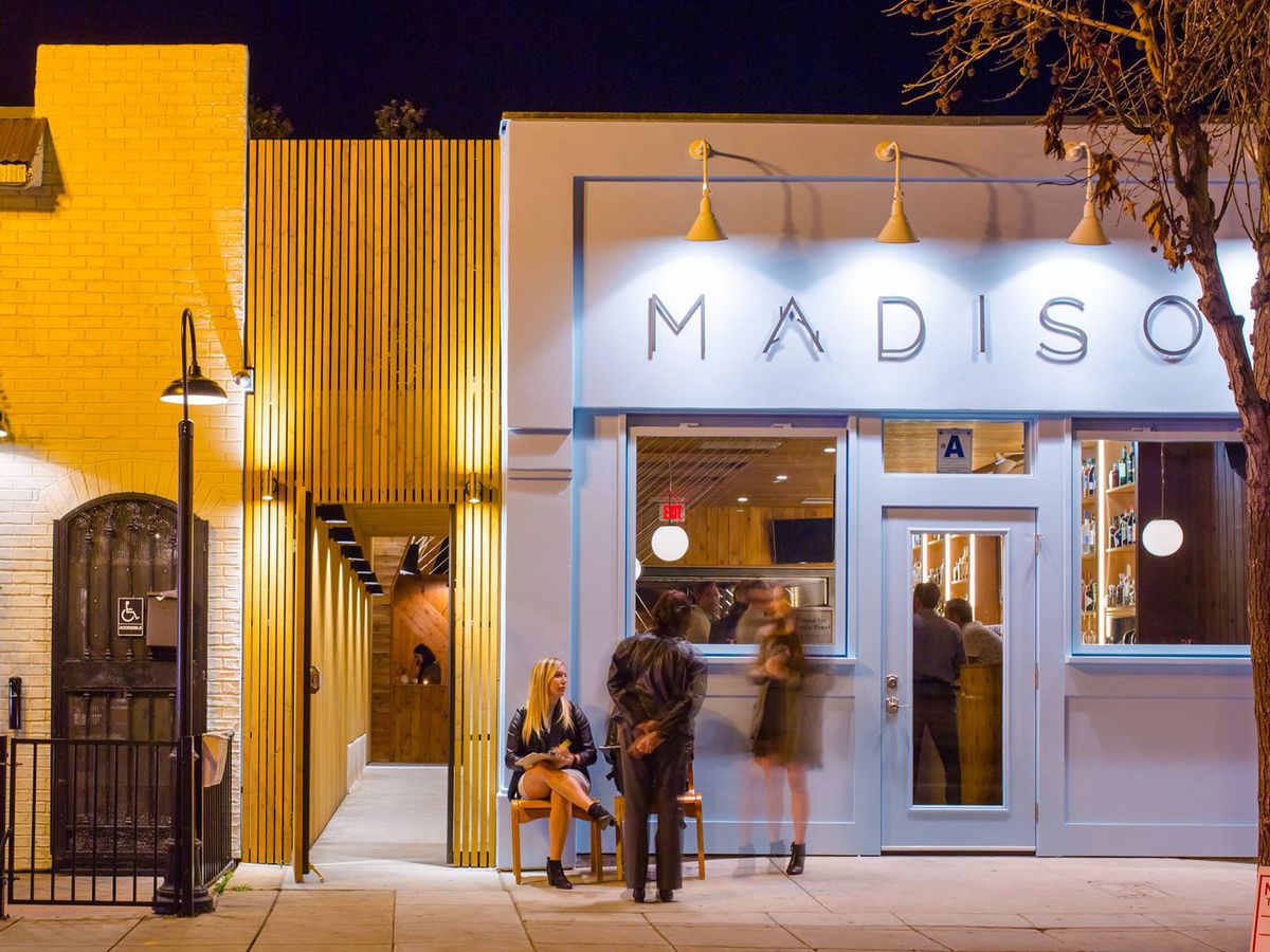 The entrance to Madison. a pastel colored building, photographed at night. Four lights brighten the restaurant’s logo, in a minimal font. A bare tree is on thje right. Several people stand outside.
