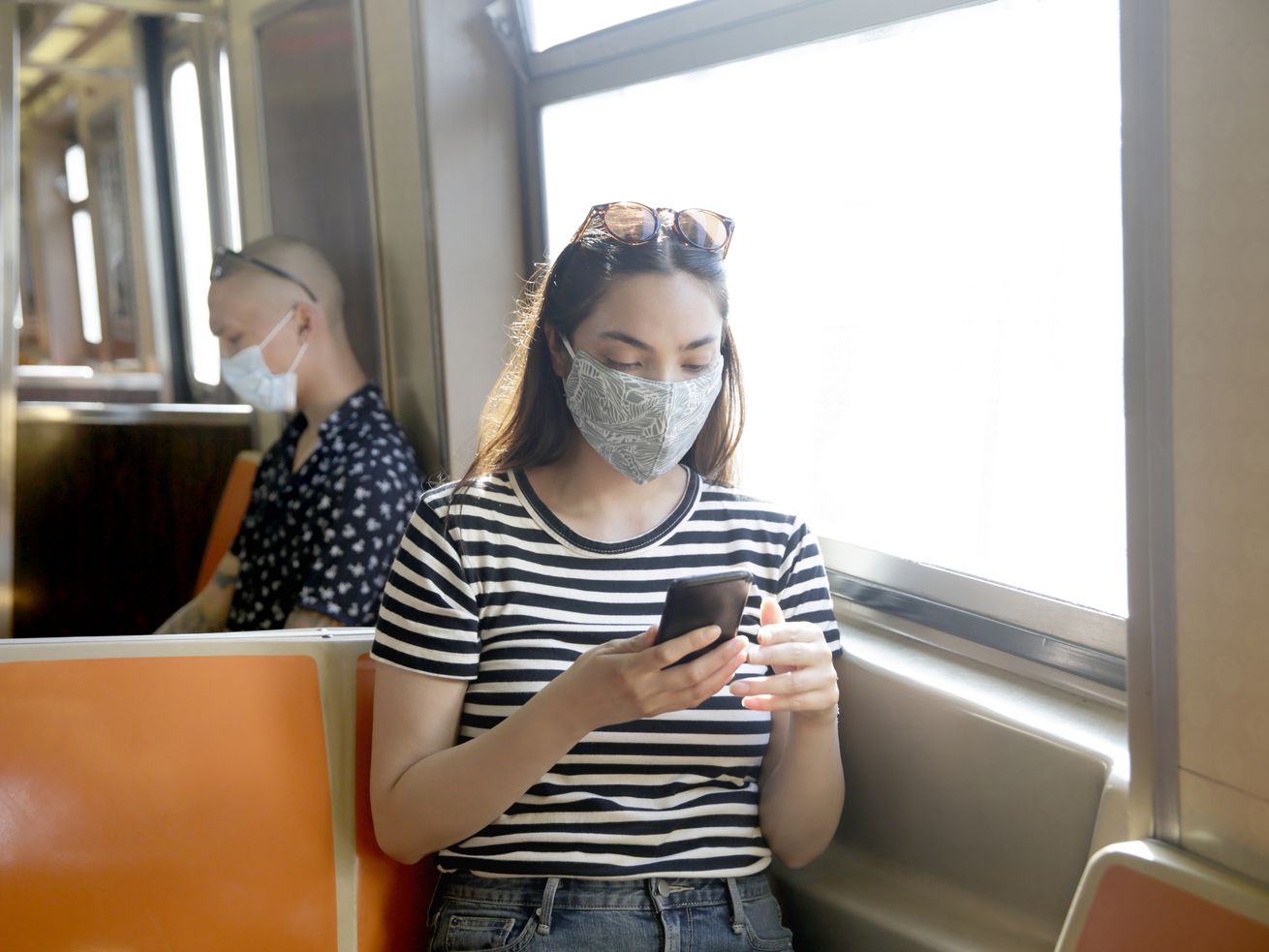 Two face-masked people sitting in a subway car, each looking at their phone.