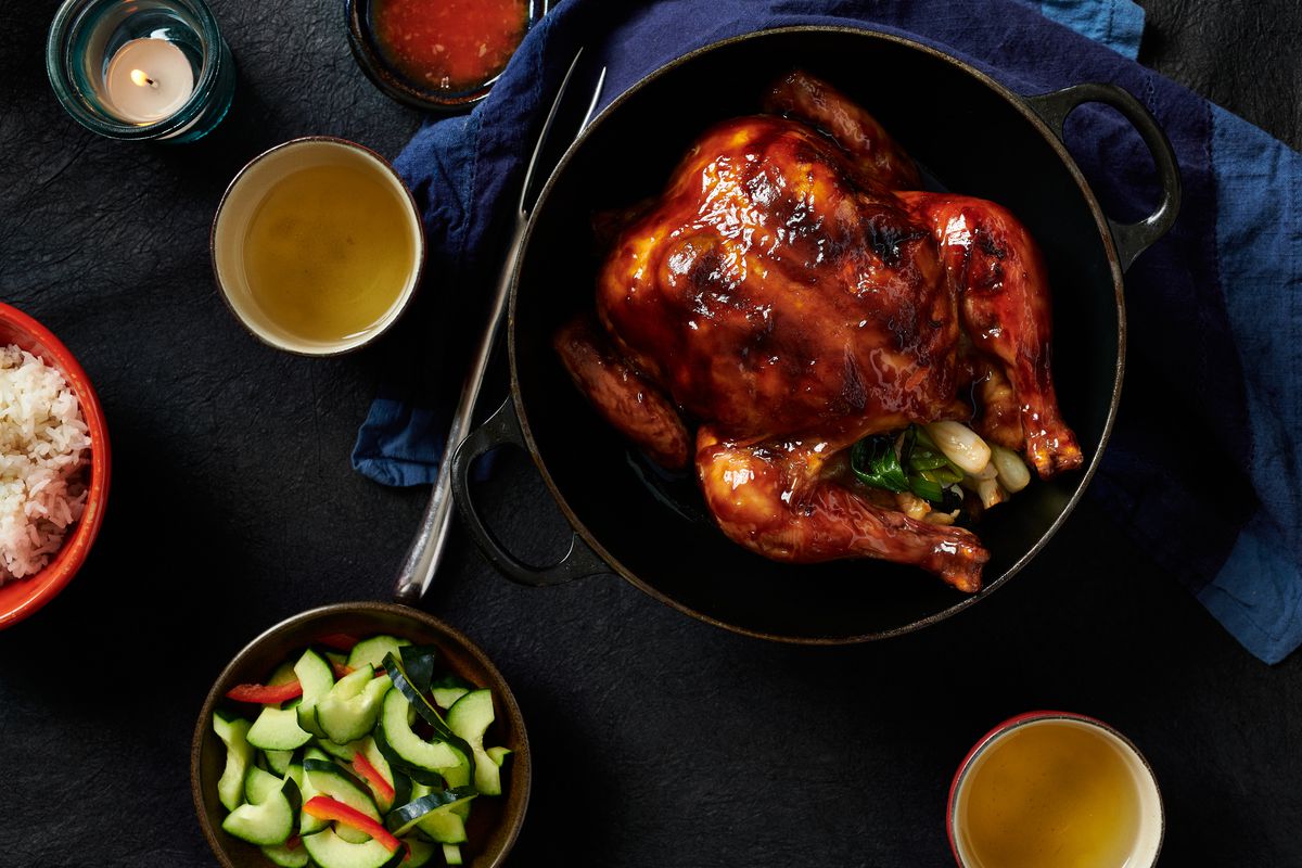 An entire chicken, its skin golden-brown, sits in a large pot on a table. Next to the pot are a small bowls of rice, sliced cucumbers, and sauce, cups of wine, and a candle.