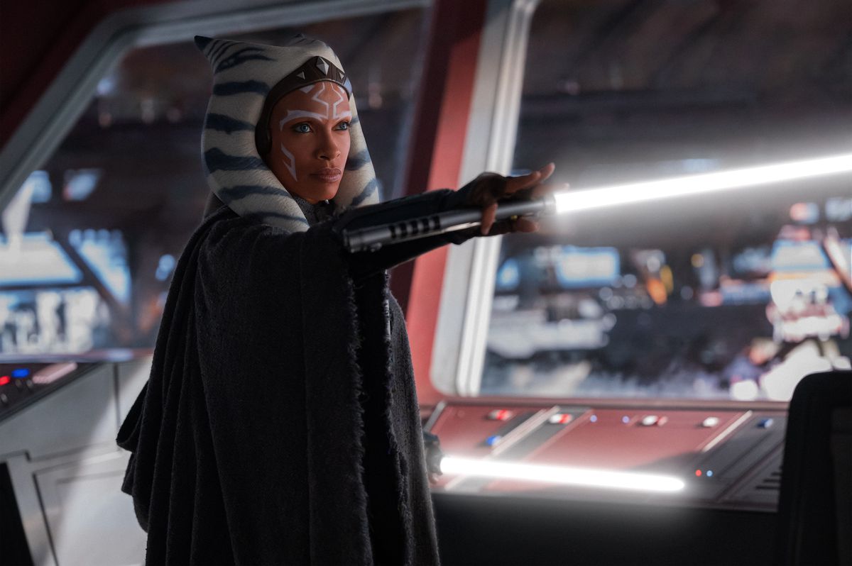 Ahsoka stands in front of a window in a cloak holding here white lightsaber horizontally in front of her in a scene from the Disney Plus series Ahsoka