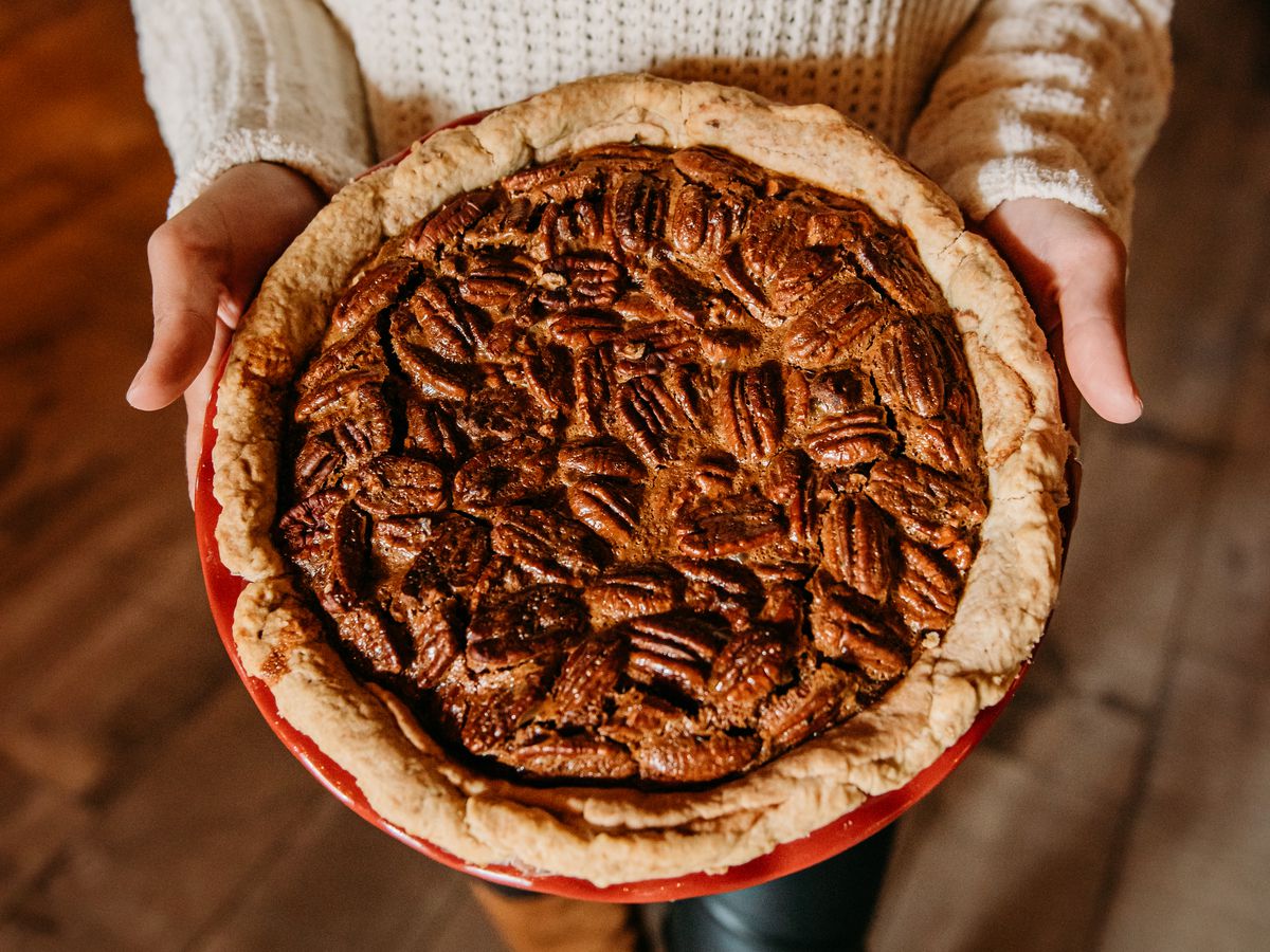 A woman in a white sweater and leather boots holding a large pie topped with pecans.