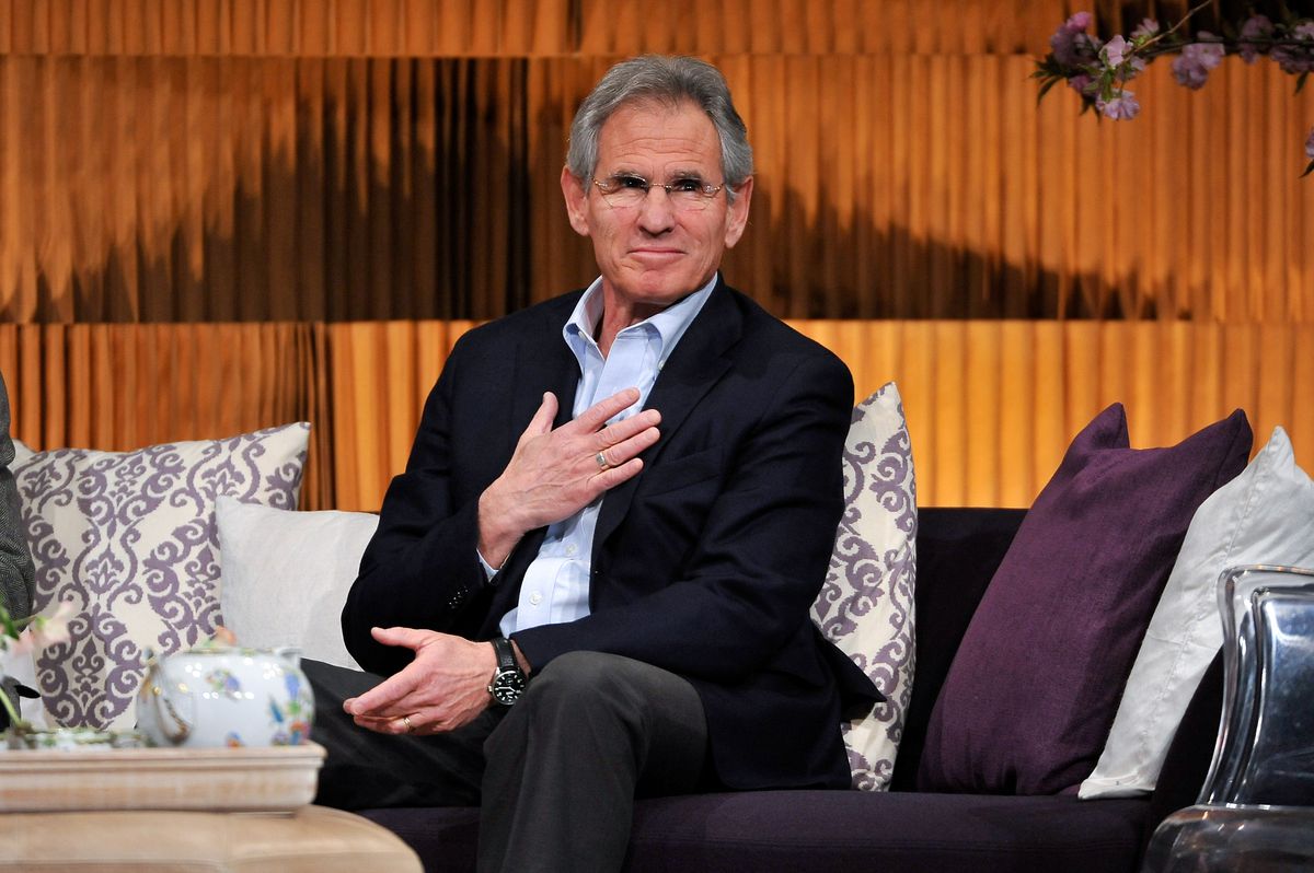 Jon Kabat-Zinn seated on a couch at the THRIVE conference, holding a hand over his heart and smiling.