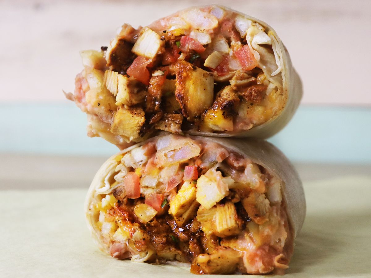 Two halves of a chicken burrito rest on a counter, overflowing with chunks of meat, refried beans, french fries, and cheese.