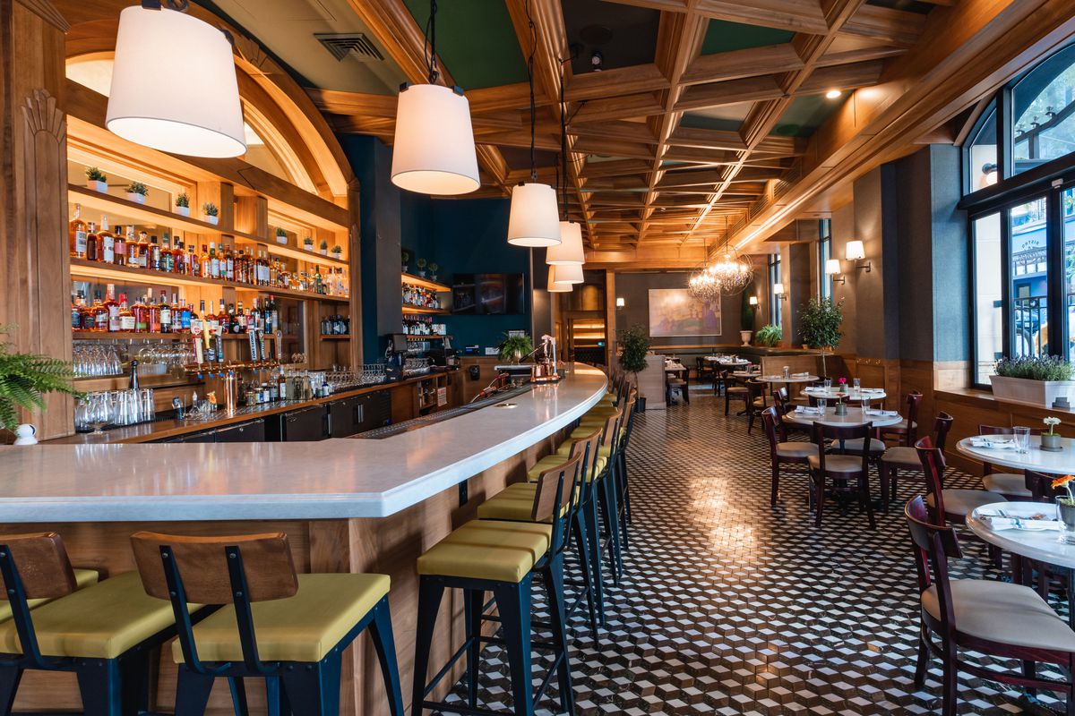 A white marble bar runs down the side of a restaurant with checkered floors and bistro tables.