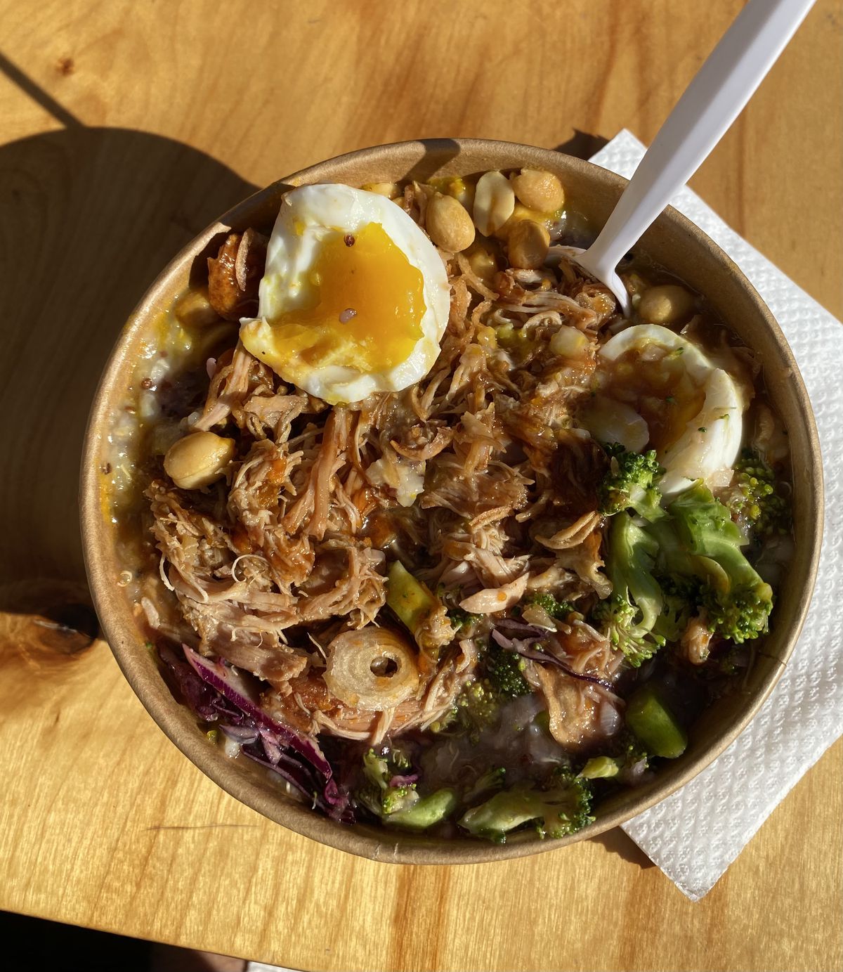 An overhead photograph of a bowl of congee, filled with soft-boiled egg, pulled chicken, broccoli, peanuts, and other ingredients