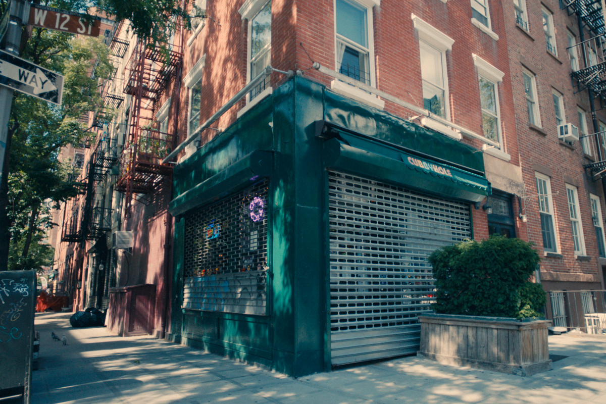A bar with green trim, neon signs, and its security gate down on a block in New York’s West Village.