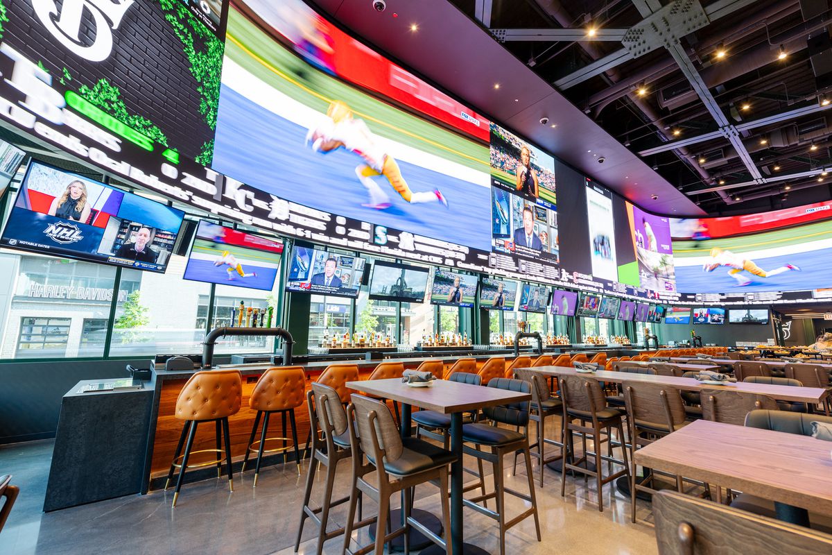 A section of a massive indoor venue with a bar and tall tables. The walls are entirely covered with TV screens of various sizes.