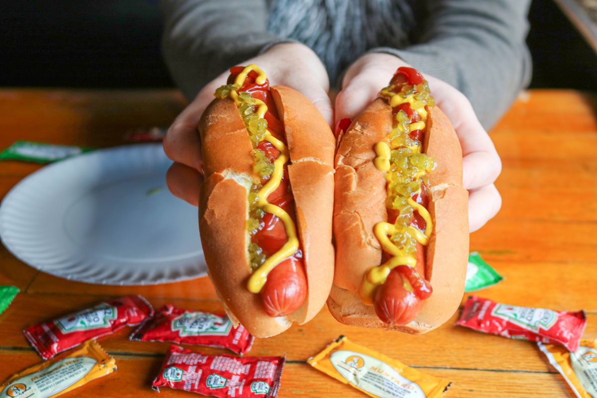 A picture of two hands holding two hot dogs with ketchup, mustard, and relish toppings.