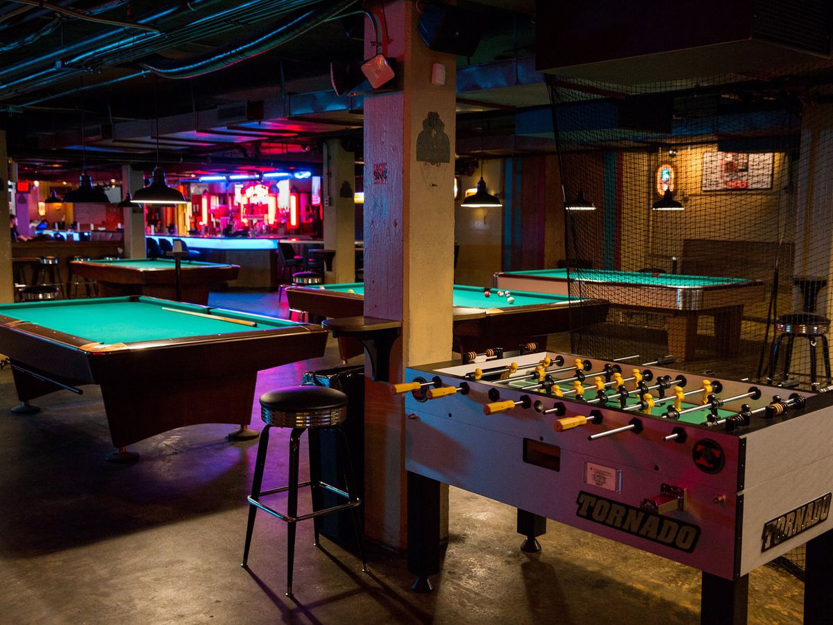 a dimly lit bar with three pool tables lined up in a row and a foosball table in front of them