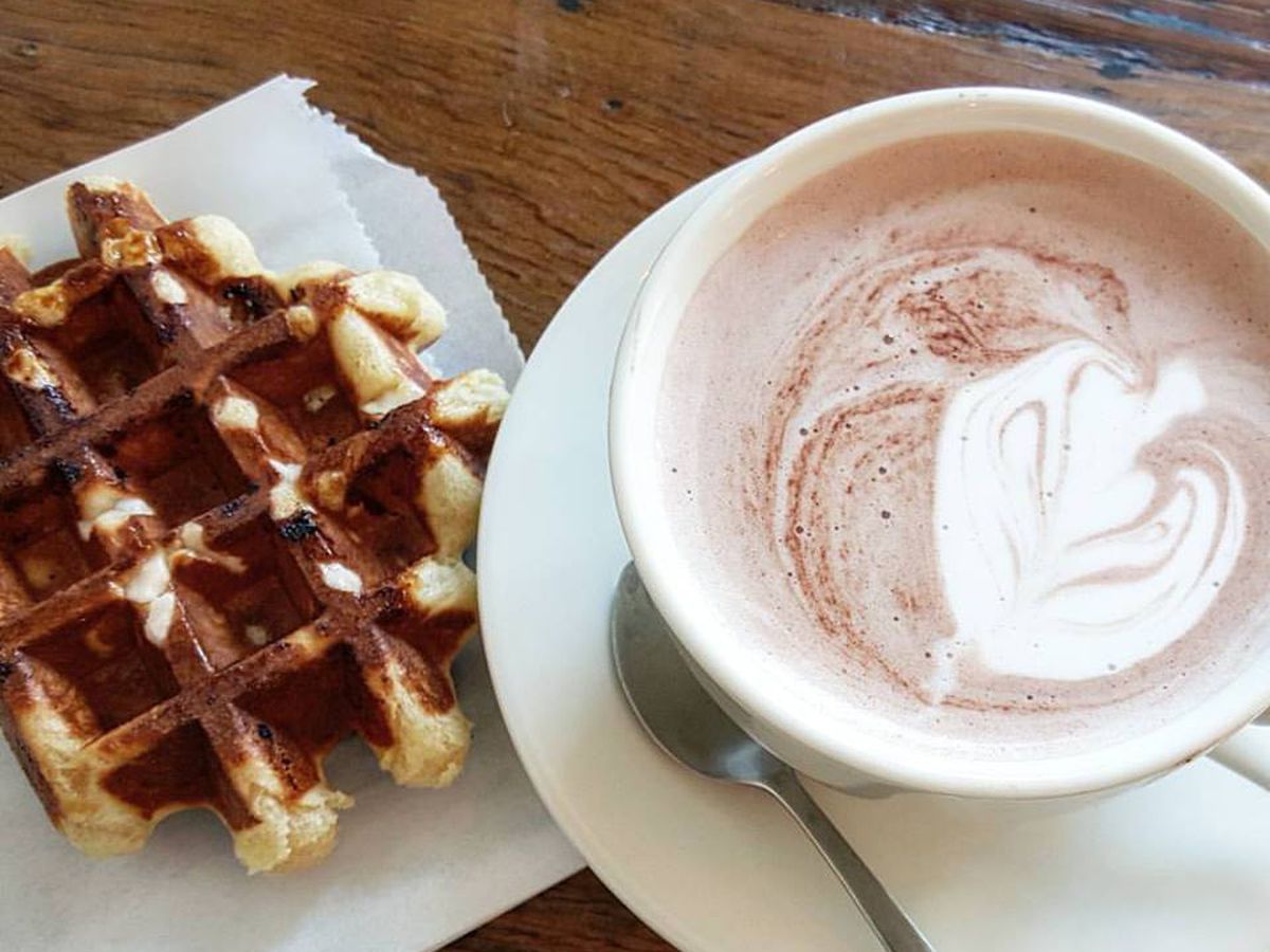 Overhead view of a liege-style waffle on a white paper bag and a white mug of hot chocolate, both on a wooden table