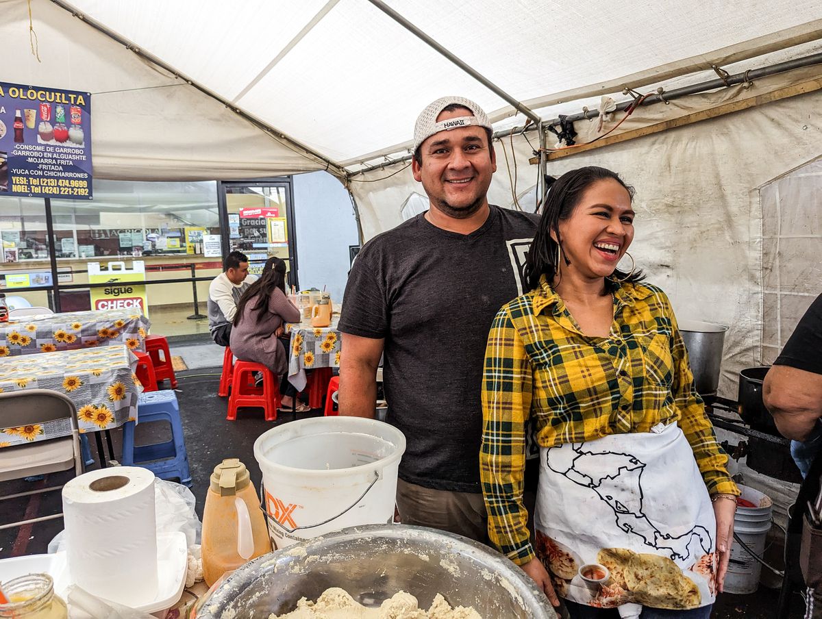 A woman in a yellow checkered shirt laughs as her partner smiles with a backwards hat on in front of bowls of food at a daytime street vendor market in Los Angeles.