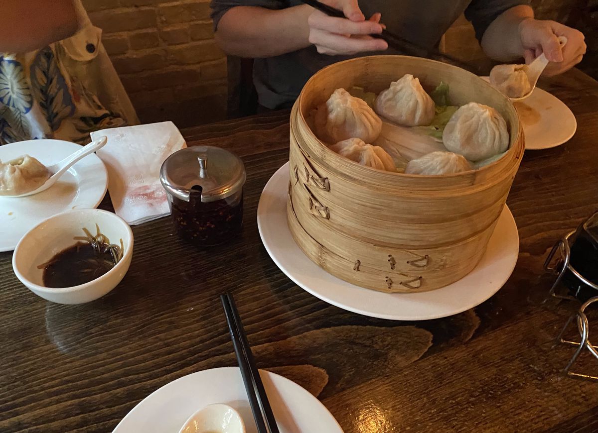 Dipping sauces and a bamboo steamer with soup dumplings are arranged on a wooden table.