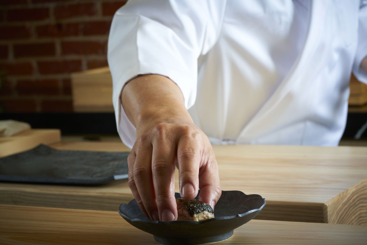 A man’s hand places a piece of sushi onto a black serving plate.