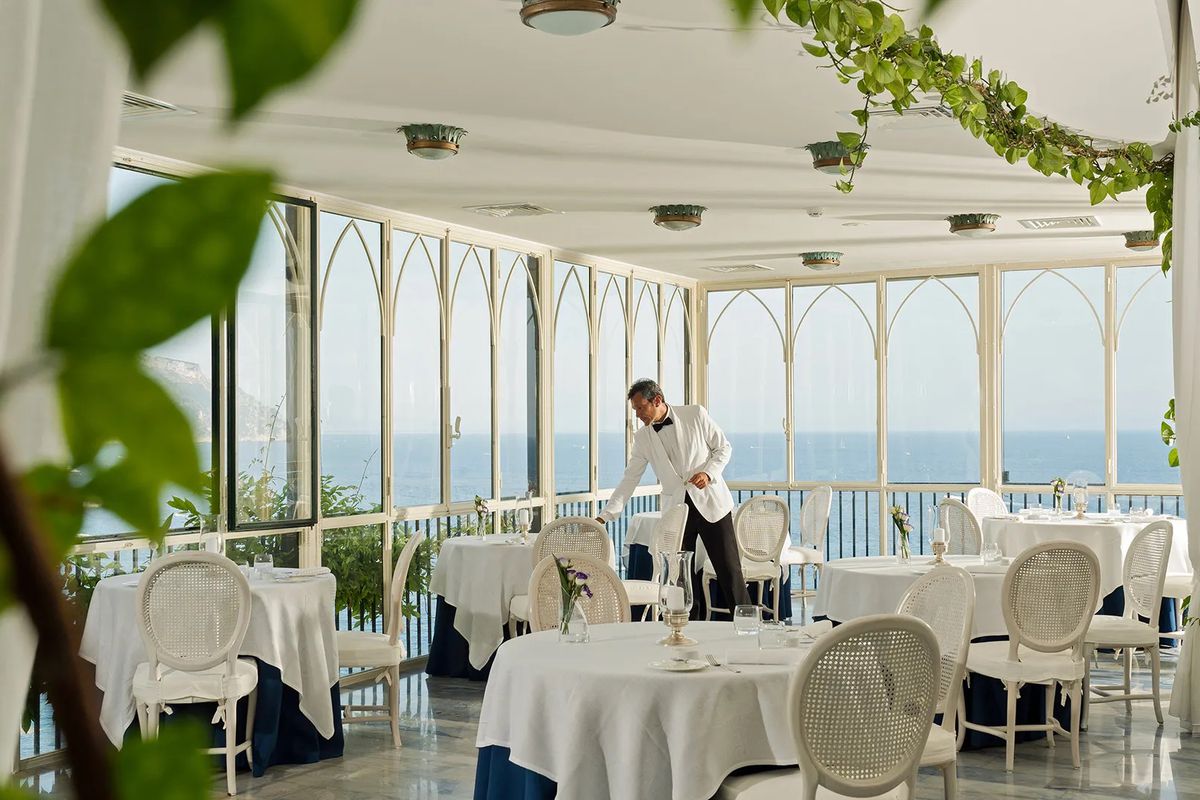 A server in a white blazer sets silverware for service in a window-wrapped coastal Italian restaurant dining room.